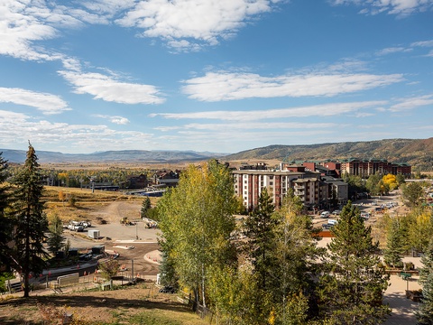 This mountain resort area features multi-story lodges surrounded by evergreen and aspen trees, with scenic hills in the background. The property is situated near local streets and parking areas.