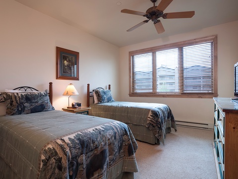 Rest easy in this cozy bedroom with two comfortable twin beds, warm lighting, and a ceiling fan to keep you cool. Your mountain getaway awaits outside those bright windows.