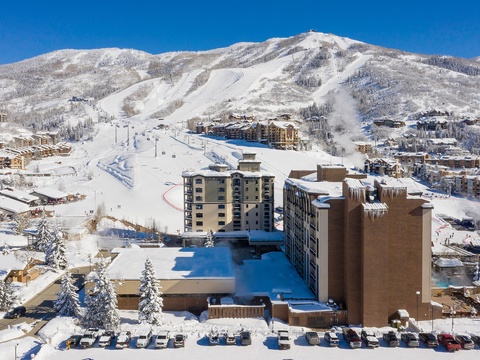 Aerial view of a premier ski resort nestled at the base of snow-covered mountain slopes, with ski lifts and trails winding through frosted evergreens under a brilliant blue winter sky.