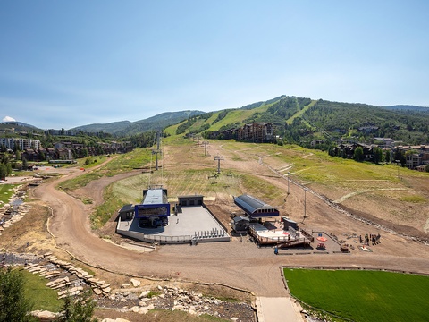 Mountain resort base area with ski slopes, summer activities venue, and surrounding lodging set among green hills under clear skies.
