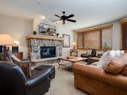 Unwind in your inviting living room with cozy leather seating, a warm stone fireplace, and mountain views through natural wood blinds creating the perfect alpine retreat.