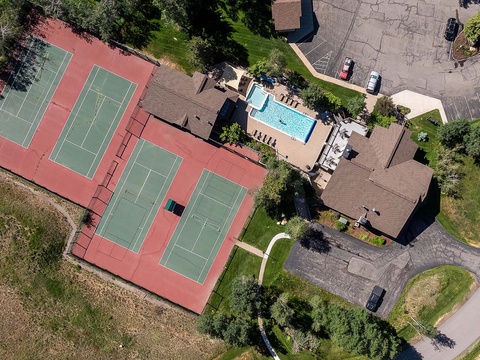 Aerial view of the property showcasing tennis courts, a sparkling pool, and ample parking surrounded by lush greenery and natural landscape.