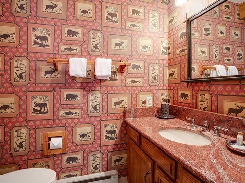 Step into this charming bathroom featuring whimsical wilderness-themed wallpaper depicting native wildlife, wooden cabinetry, and warm granite countertops that create a cozy cabin retreat atmosphere.