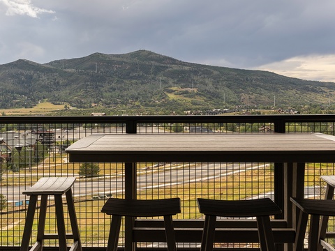 Scenic mountain landscape viewed from a covered balcony or terrace area with wire railing and seating table, featuring rolling hills and valley terrain.