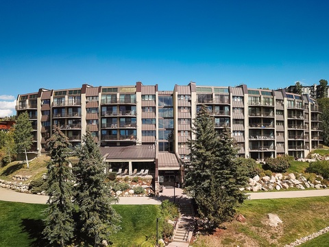A multi-story mountain resort building with spacious balconies, nestled in a scenic alpine setting with manicured lawns, evergreen trees, and natural stone landscaping.
