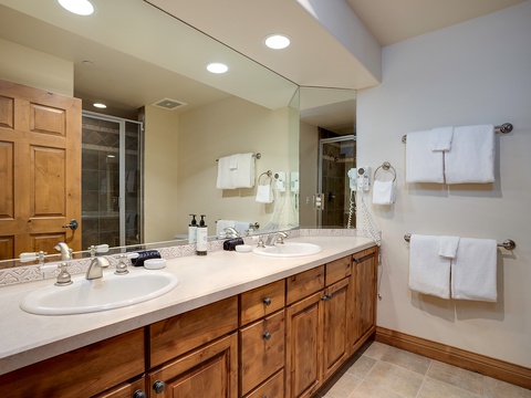 Step into your private spa-like retreat with dual vanities, warm wood cabinetry, plush towels, and a spacious glass-enclosed shower waiting just for you.