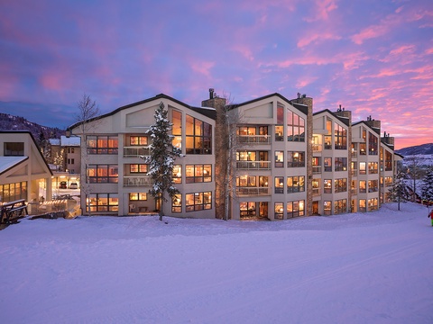 Modern mountain property nestled in a snowy alpine setting, illuminated at dusk with striking sunset skies painting the horizon in vibrant pink and purple hues.