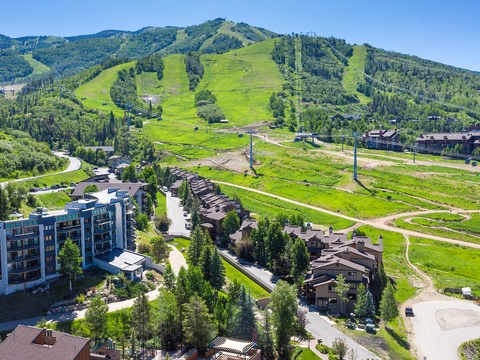Aerial view of a mountain resort community nestled at the base of summer ski slopes, surrounded by lush green meadows and forested peaks.