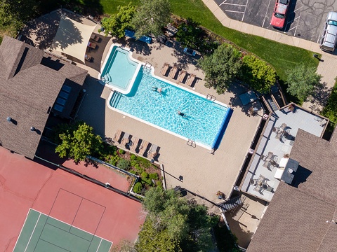 Aerial view of the property featuring a sparkling outdoor swimming pool surrounded by lounge chairs, tennis court, and landscaped green spaces with ample parking nearby.