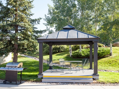 A covered pavilion with dining seating and a BBQ grill nestled in landscaped grounds, perfect for relaxing outdoor meals surrounded by evergreens and rolling lawns.