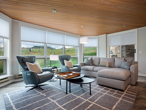 Unwind in your spacious living room with floor-to-ceiling windows framing peaceful meadow views. Sink into plush seating and let the natural light and warm wood ceiling create your perfect retreat.