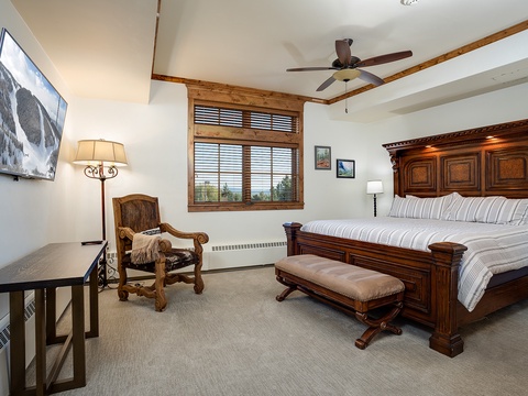 Unwind in your spacious bedroom featuring a handcrafted bed, cozy seating area, and views of the surrounding landscape through rustic wooden blinds. Entertainment awaits on the large flat-screen TV.
