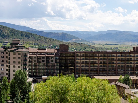 Multi-story resort building set against scenic mountain terrain with forested hillsides and a valley floor visible in the background, surrounded by trees and offering elevated views of the landscape.