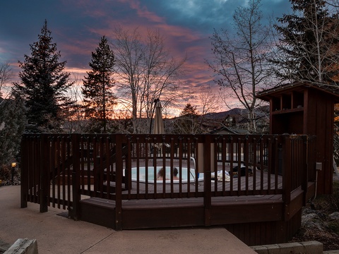 Property features a hot tub area surrounded by wooden railings, set against a backdrop of evergreen trees and colorful twilight sky.