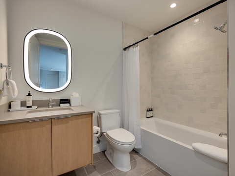 Step into your spa-like bathroom with its glowing backlit mirror, spacious tub, and rainfall shower. Soft white tiles and warm wood vanity create a serene space perfect for unwinding after your day.