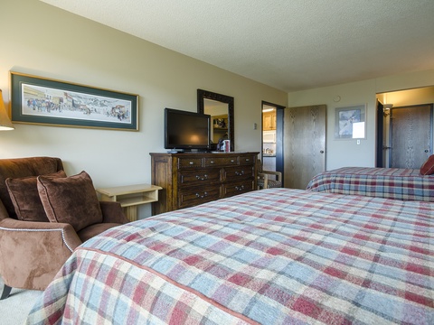 Your spacious bedroom features two cozy plaid-covered beds, a plush armchair perfect for reading, and a flat-screen TV for relaxing evenings. Rustic charm meets comfort in every detail.