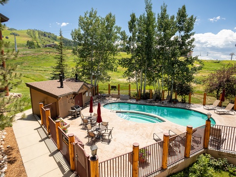 Scenic mountain resort pool area nestled among rolling green hills, with ski lift access visible in the distance and mature trees providing natural shade throughout the seasons.