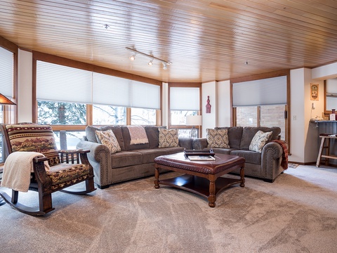 Unwind in your inviting living room with plush sectional seating, a cozy rocking chair, and warm wood-paneled ceilings that frame beautiful winter views just beyond your windows.