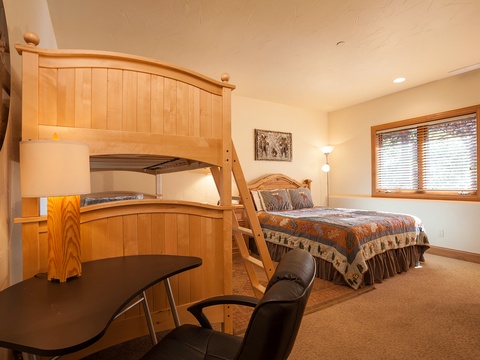 Your cozy retreat features a handcrafted wooden bunk bed and a comfortable queen bed dressed in rustic linens. Natural light streams through the window, illuminating warm wood tones and thoughtful furnishings for relaxation.
