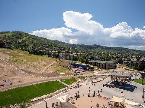 Aerial view of a mountain resort community nestled in rolling hills with diverse residential buildings, outdoor recreation areas, and scenic mountain backdrops under dramatic summer clouds.