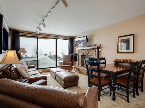 Cozy living and dining space where you'll unwind after mountain adventures, with snowy views beyond sliding glass doors, a stone fireplace for warmth, and inviting leather seating.
