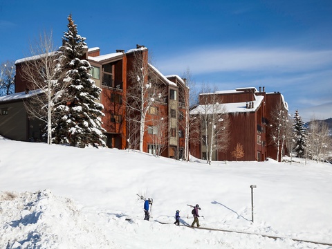 Mountain vacation rental complex featuring rustic wood and stone architecture nestled in a snowy alpine setting, with ski-in access and surrounded by evergreens.