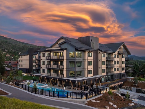 A modern mountain resort property showcasing contemporary architecture with dramatic cloud formations overhead, nestled among rolling hillsides and neighboring structures in the surrounding valley.