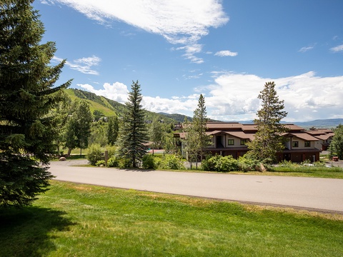Mountain resort property nestled in a scenic valley, surrounded by evergreen trees and framed by distant peaks under a blue summer sky.