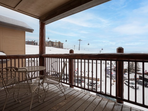 A covered balcony overlooking a snowy ski village provides a perfect spot to watch skiers glide down the slopes, with ski lifts visible in the distance and cozy seating for your morning coffee.