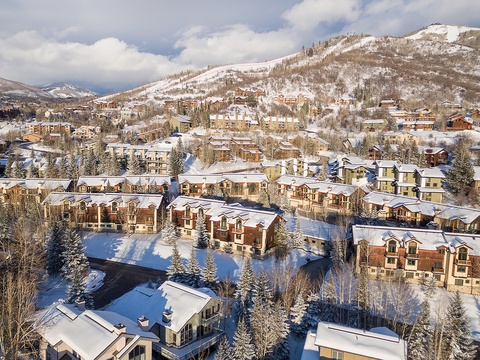 Aerial perspective of a snow-covered mountain resort community, showcasing vacation rental properties nestled among evergreen trees with ski slopes and peaks rising in the distance.