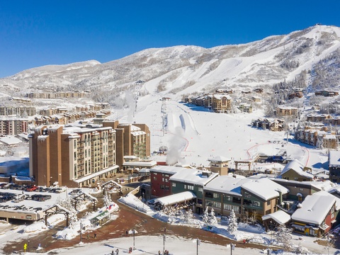 Aerial view of a premier ski resort village nestled in snow-covered mountains, featuring ski-in/ski-out access, ski lifts, and alpine lodges under clear blue skies.