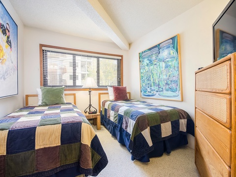 Cozy twin beds invite restful nights beneath vibrant artwork and natural light filtering through the window, with ample storage in the warm wood dresser by your side.