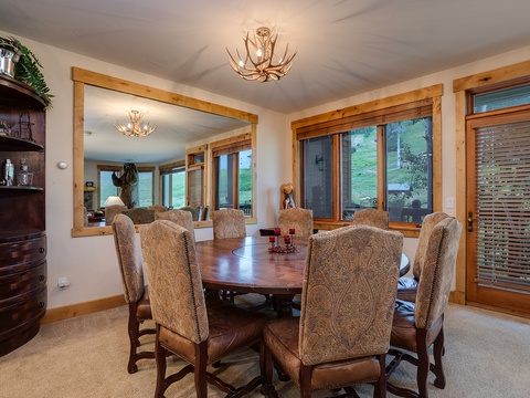 Gather around this elegant dining table, where rustic antler chandeliers cast a warm glow and large windows frame peaceful views, making every meal feel special in your mountain retreat.