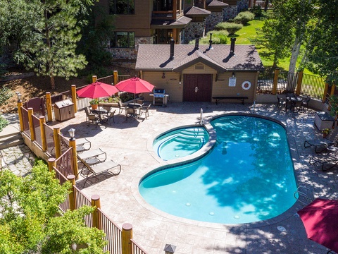 Aerial view of the property featuring a private pool, hot tub, and patio dining area with BBQ facilities, surrounded by lush trees and mountain greenery.
