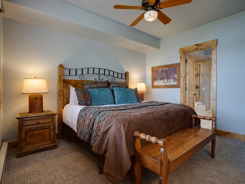 Your peaceful retreat features a plush bed with inviting linens, warm bedside lighting, and a ceiling fan for comfort. The ensuite bath awaits just steps away through rustic wood doors.