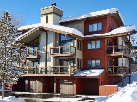 Modern ski chalet nestled in snowy mountain landscape, featuring warm red cedar siding, multiple balconies, and inviting covered entry with chimney rising above snow-capped roofline.