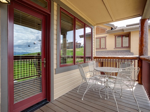 A covered deck with seating for four overlooks rolling green hills, nearby buildings, and distant mountain views—perfect for morning coffee or evening relaxation in the fresh mountain air.