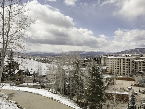 Scenic winter mountain resort area featuring snow-dusted peaks, evergreen forests, and multi-story lodgings nestled in a valley under dramatic cloudy skies.
