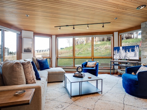Sink into plush seating surrounded by floor-to-ceiling windows framing vibrant hillside views. The warm wood ceiling and modern fireplace create your perfect retreat for mountain relaxation.