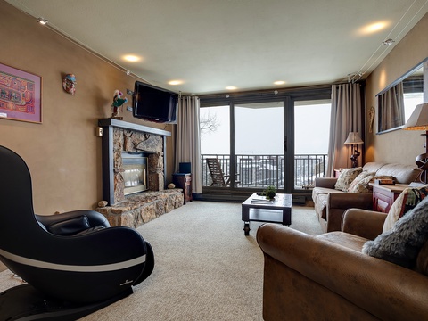 Your inviting living space features plush sofas, a cozy stone fireplace, and a private balcony where you can unwind while enjoying scenic mountain views through floor-to-ceiling windows.