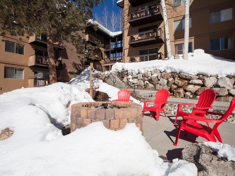 The property sits in a winter mountain setting with bright red seating around an outdoor fire pit, snow-covered stone walls, and multi-story buildings nearby.