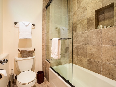 Your private bathroom features warm stone tile, a glass-enclosed tub-shower combo, and crisp white towels ready for your arrival.