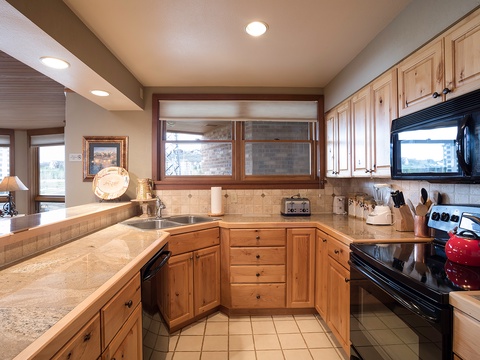 This fully-equipped kitchen features natural wood cabinetry, tiled countertops, and modern black appliances. Windows frame city views, while thoughtful touches like the red kettle add homey warmth to your cooking space.