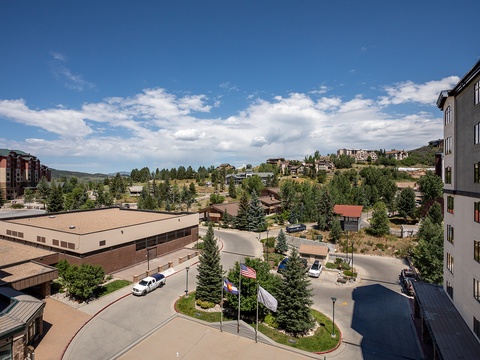 Mountain resort setting with nearby residential buildings, lush evergreens, and clear summer skies. The property is located in a scenic hillside community with convenient access to local amenities.