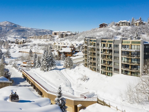 This alpine village resort nestled in snow-covered mountains offers stunning winter scenery. Modern multi-story buildings sit amid snowy pines, with ski lodges dotting the mountainside under clear blue skies.