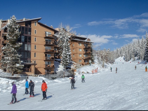 Multi-story mountain resort buildings nestled at the base of ski slopes, offering convenient ski-in/ski-out access surrounded by snow-covered evergreens under blue skies.