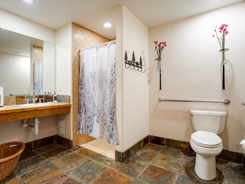 Your spacious bathroom features a walk-in shower with forest-themed curtain, warm tile flooring, and thoughtful grab bars for comfort and safety. Fresh flowers add a welcoming touch to your mountain retreat.