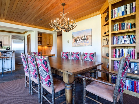Gather around this rustic wood dining table beneath a striking antler chandelier, with built-in bookshelves and colorful patterned seating inviting your group to share meals and stories.