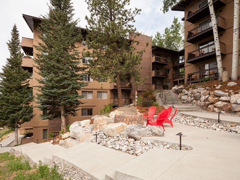The property features multi-story residential buildings nestled among evergreens, with landscaped outdoor spaces including a fire pit and red Adirondack chairs for guests to enjoy the mountain surroundings.