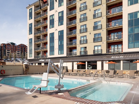 The property features a modern multi-level design with private balconies and an outdoor pool area complete with lounge seating and covered dining space, set against a scenic mountain backdrop.